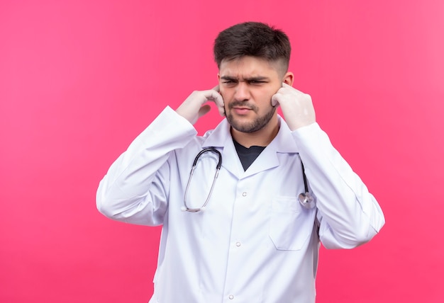 Giovane medico bello che porta guanti medici bianchi dell'abito medico bianco e stetoscopio che chiude le sue orecchie con le mani in piedi sopra la parete rosa