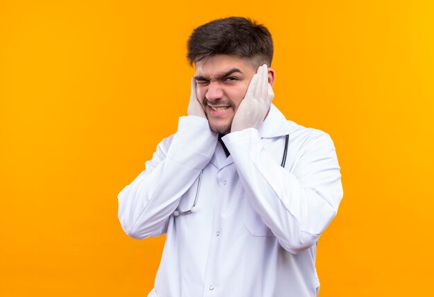 Giovane medico bello che porta guanti medici bianchi e stetoscopio bianchi dell'abito medico che si lamentano del suono forte che sta sopra la parete arancione