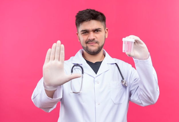 Giovane medico bello che porta guanti medici bianchi e stetoscopio bianchi dell'abito medico, che tiene il contenitore trasparente di analisi che fa il segnale di stop con la mano che sta sopra la parete rosa