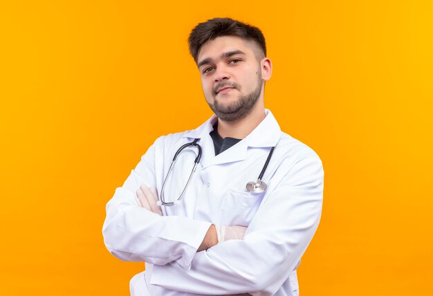 Giovane medico bello che porta i guanti medici bianchi dell'abito medico bianco e lo stetoscopio che sembrano smudgly che si levano in piedi sopra la parete arancione