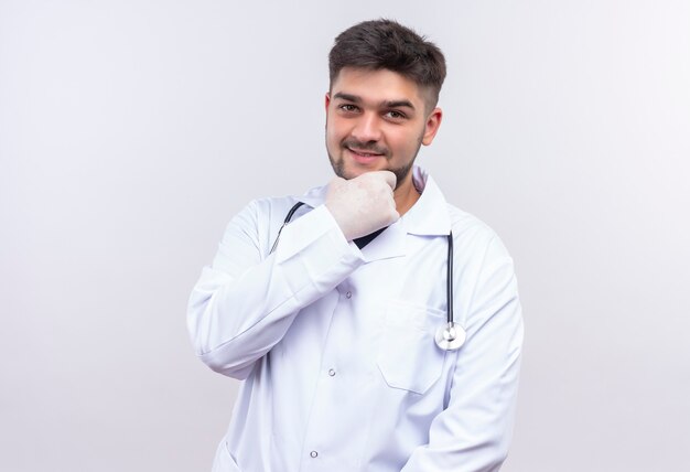 Giovane medico bello che porta i guanti medici bianchi dell'abito medico bianco e lo stetoscopio che tiene la mascella con la mano e sorridere