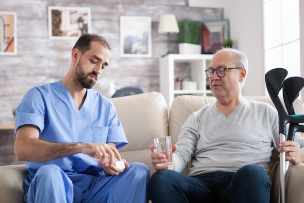 Giovane medico caucasico in casa di cura che dà le pillole all'uomo anziano con le stampelle. .