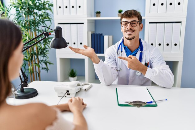 Giovane medico con cliente presso la clinica medica stupito e sorridente alla telecamera mentre si presenta con la mano e punta con il dito.