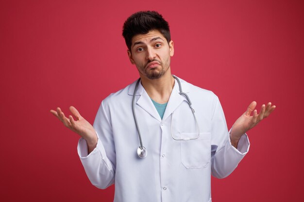 Giovane medico di sesso maschile senza indizi che indossa uniforme medica e stetoscopio intorno al collo guardando la fotocamera che mostra che non conosco il gesto isolato su sfondo rosso