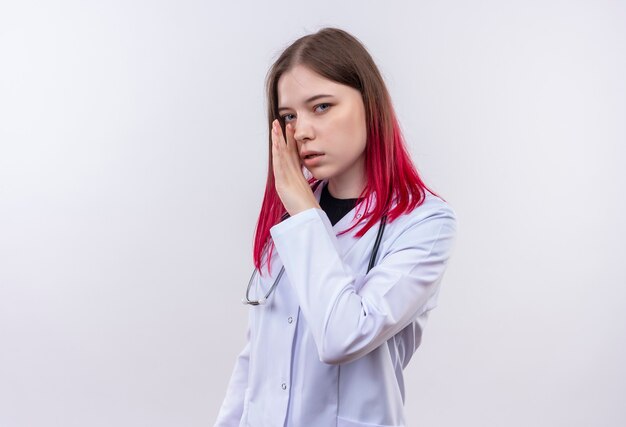 giovane medico donna che indossa uno stetoscopio sussurra veste medica sul muro bianco isolato