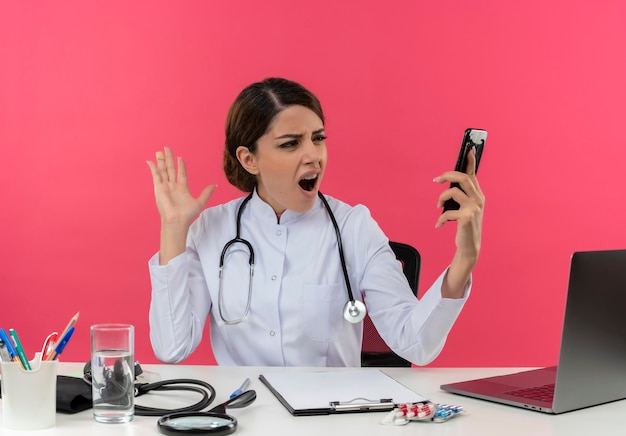 Giovane medico femminile arrabbiato che indossa veste medica e stetoscopio seduto alla scrivania con strumenti medici e laptop tenendo e guardando il telefono cellulare tenendo la mano in aria isolata sul muro rosa