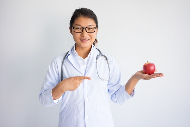 Giovane medico femminile asiatico sorridente che tiene e che indica alla mela rossa.