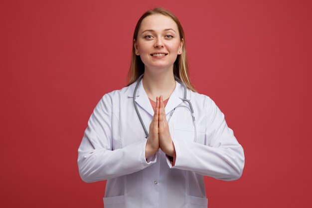 Giovane medico femminile biondo sorridente che porta veste medica e stetoscopio intorno al collo che fa gesto di namaste