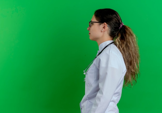 Giovane medico femminile che indossa veste medica e stetoscopio e occhiali in piedi in vista di profilo guardando dritto isolato sulla parete verde con lo spazio della copia