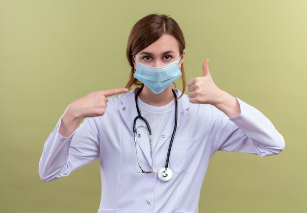 Giovane medico femminile che indossa veste medica, maschera e stetoscopio che mostra il pollice in su e indicandolo sulla parete verde isolata