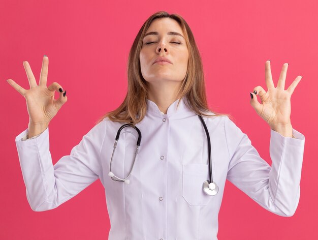 Giovane medico femminile con gli occhi chiusi che indossa la veste medica con lo stetoscopio che tiene le pillole che mostrano il gesto di meditazione isolato sulla parete rosa