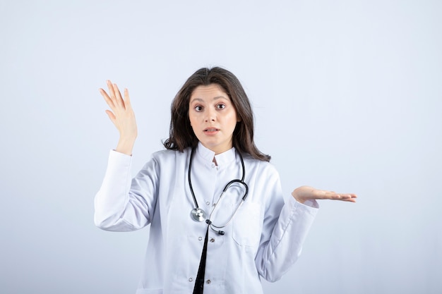 Giovane medico femminile con lo stetoscopio che propone alla macchina fotografica sul muro bianco.