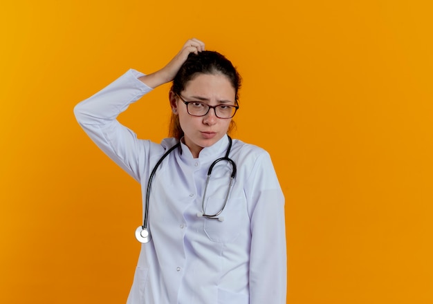 Giovane medico femminile confuso che indossa veste medica e stetoscopio con gli occhiali che graffiano la testa isolata