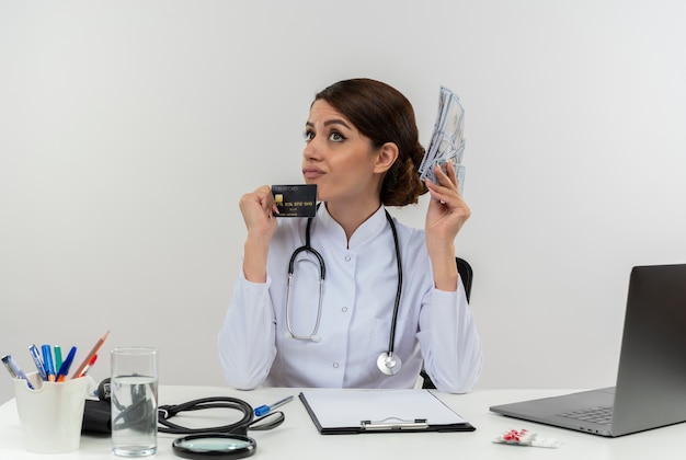 Giovane medico femminile di pensiero che indossa la veste medica con lo stetoscopio che si siede alla scrivania lavora sul computer con strumenti medici che tengono contanti e carta di credito con lo spazio della copia