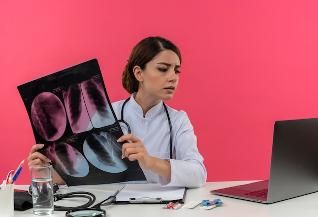 Giovane medico femminile di pensiero che indossa la veste medica con lo stetoscopio che si siede alla scrivania lavora sul computer con strumenti medici che tengono i raggi x e guardando il computer portatile con lo spazio della copia