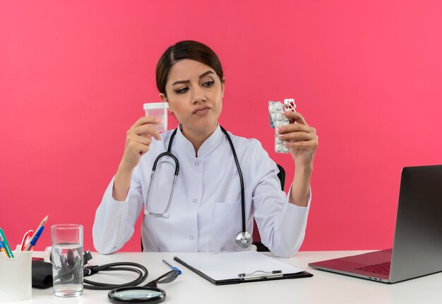 Giovane medico femminile di pensiero che indossa la veste medica con lo stetoscopio che si siede alla scrivania lavora sul computer con strumenti medici che tengono lattina vuota e pillole con lo spazio della copia