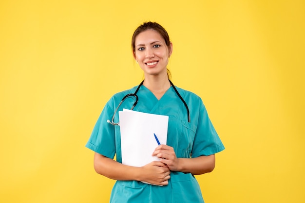 Giovane medico femminile di vista frontale in camicia medica che tiene analisi della carta su fondo giallo