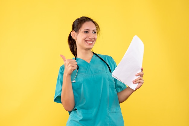 Giovane medico femminile di vista frontale in camicia medica che tiene analisi della carta su fondo giallo