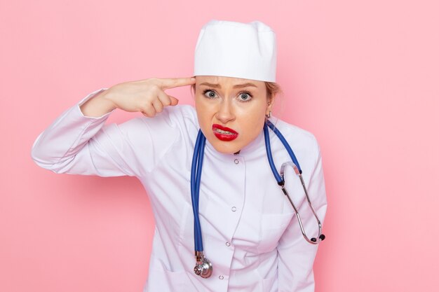 Giovane medico femminile di vista frontale in vestito bianco con lo stetoscopio blu che posa con l'espressione divertente sul lavoro femminile medico dell'ospedale medico dello spazio rosa