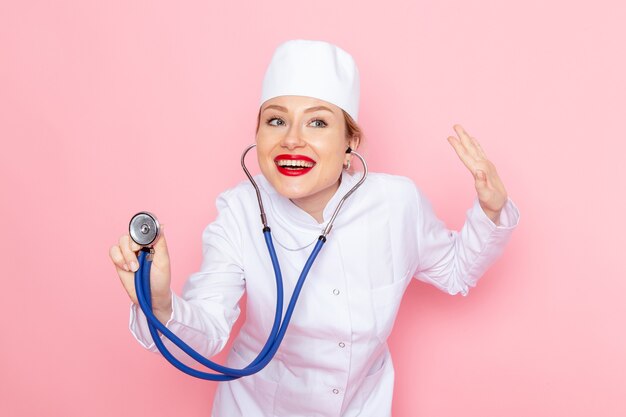 Giovane medico femminile di vista frontale in vestito bianco con lo stetoscopio blu che posa sorridente e che misura sul lavoro femminile medico dell'ospedale medico della medicina dello spazio rosa