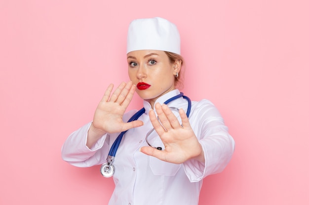 Giovane medico femminile di vista frontale in vestito bianco con lo stetoscopio blu che posa sul lavoro rosa dello spazio
