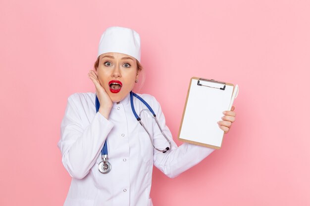 Giovane medico femminile di vista frontale in vestito medico bianco con il blocchetto per appunti blu della tenuta dello stetoscopio che posa sull'ospedale medico rosa della medicina dello spazio