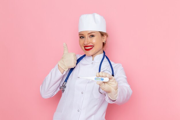 Giovane medico femminile di vista frontale in vestito medico bianco con il dispositivo blu della tenuta dello stetoscopio con il sorriso sull'ospedale medico rosa della medicina dello spazio