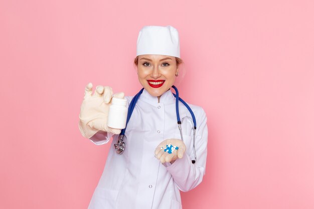 Giovane medico femminile di vista frontale in vestito medico bianco con le pillole blu della tenuta dello stetoscopio con il sorriso sullo spazio rosa