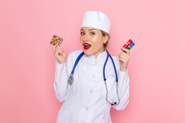 Giovane medico femminile di vista frontale in vestito medico bianco con le pillole e le boccette blu della tenuta dello stetoscopio sull'ospedale medico rosa della medicina dello spazio