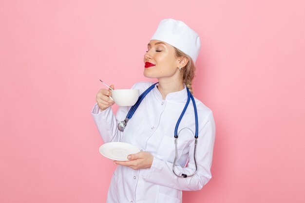 Giovane medico femminile di vista frontale in vestito medico bianco con lo stetoscopio blu che beve caffè sul lavoro medico dell'ospedale della medicina dello spazio rosa