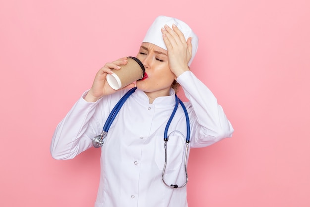 Giovane medico femminile di vista frontale in vestito medico bianco con lo stetoscopio blu che beve caffè sull'ospedale medico rosa della medicina dello spazio