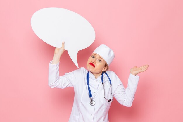 Giovane medico femminile di vista frontale in vestito medico bianco con lo stetoscopio blu che tiene il segno bianco enorme sull'infermiera medica dell'ospedale di medicina dello spazio rosa