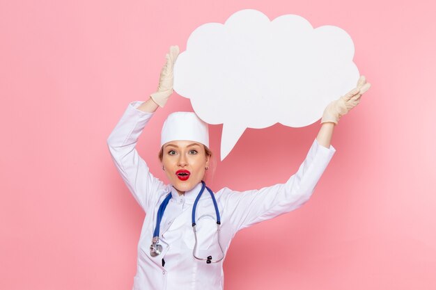 Giovane medico femminile di vista frontale in vestito medico bianco con lo stetoscopio blu che tiene il segno bianco enorme sulla salute medica dell'ospedale medico della medicina dello spazio rosa