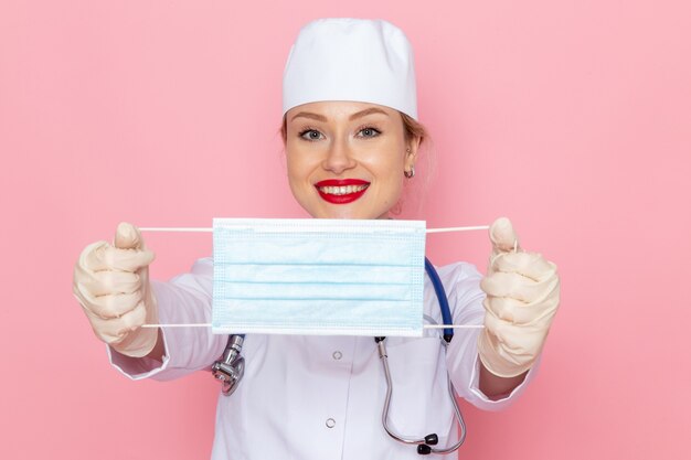 Giovane medico femminile di vista frontale in vestito medico bianco con lo stetoscopio blu che tiene maschera sterile sull'infermiera rosa dello spazio