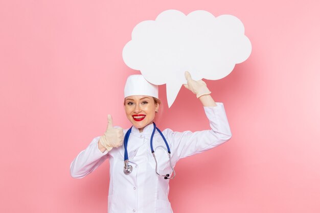 Giovane medico femminile di vista frontale in vestito medico bianco con lo stetoscopio blu che tiene segno bianco enorme con il sorriso sul lavoro di salute dell'ospedale medico di medicina dello spazio rosa