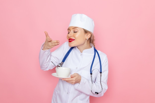 Giovane medico femminile di vista frontale in vestito medico bianco con lo stetoscopio blu che tiene tazza di caffè sull'ospedale medico della medicina dello spazio rosa