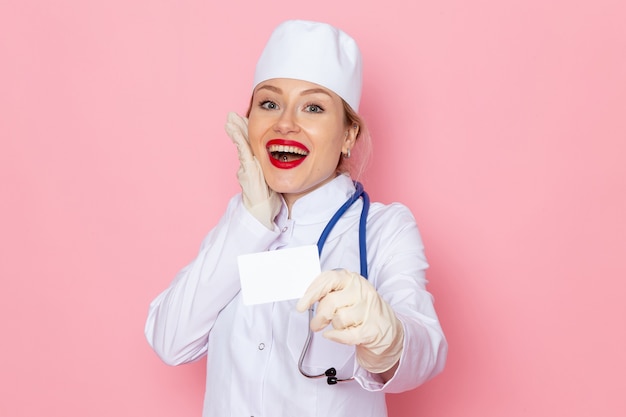 Giovane medico femminile di vista frontale in vestito medico bianco con stetoscopio blu che tiene carta bianca che sorride sull'infermiera dell'ospedale medico della medicina dello spazio rosa