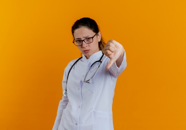Giovane medico femminile dispiaciuto che indossa abito medico e stetoscopio con gli occhiali che mostrano il pollice verso il basso isolato