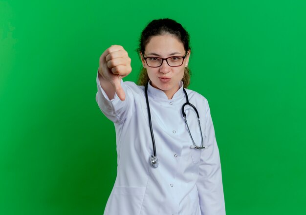 Giovane medico femminile dispiaciuto che indossa veste medica e stetoscopio e occhiali che mostrano il pollice verso il basso isolato sulla parete verde con lo spazio della copia