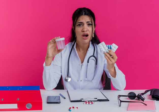 Giovane medico femminile dispiaciuto che indossa veste medica e stetoscopio seduto alla scrivania con strumenti medici che tengono farmaci e becher isolato sulla parete rosa