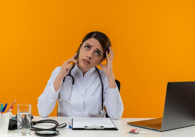 Giovane medico femminile dispiaciuto che indossa veste medica e stetoscopio seduto alla scrivania con strumenti medici e laptop parlando al telefono testa commovente cercando isolato