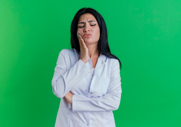 Giovane medico femminile dolorante che indossa veste medica mettendo la mano sulla guancia che soffre di mal di denti con gli occhi chiusi isolato sulla parete verde con lo spazio della copia