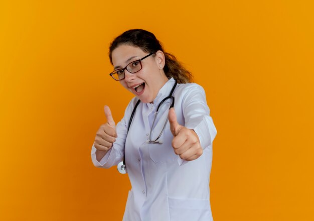 Giovane medico femminile impressionato che porta veste e stetoscopio e vetri medici che sembrano mostrare i pollici in su isolati