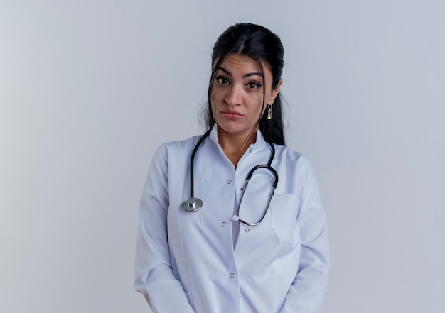Giovane medico femminile impressionato che porta veste medica e stetoscopio che sembrano isolati