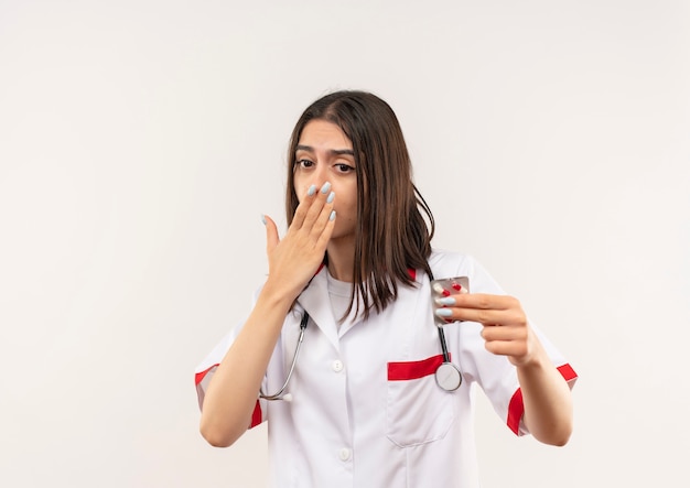 Giovane medico femminile in camice bianco con lo stetoscopio intorno al collo che mostra il blister con le pillole che sembra sorpreso e confuso che copre la bocca con la mano che sta sopra il muro bianco