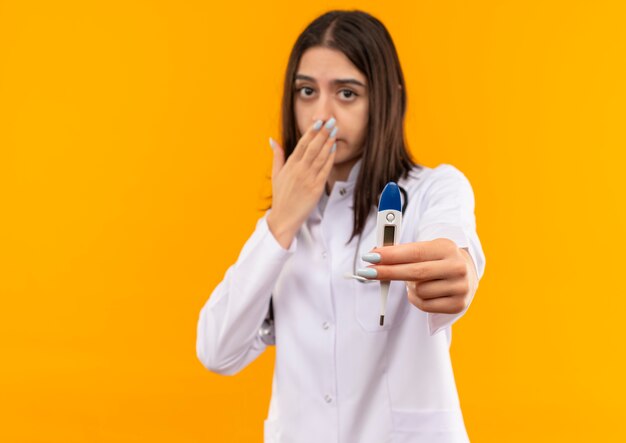 Giovane medico femminile in camice bianco con lo stetoscopio intorno al collo che mostra la bocca scioccata della copertura del termometro digitale con la mano che sta sopra la parete arancione