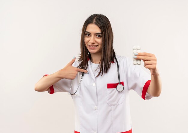 Giovane medico femminile in camice bianco con lo stetoscopio intorno al collo che mostra la bolla con le pillole che punta con il dito ad esso in piedi sul muro bianco