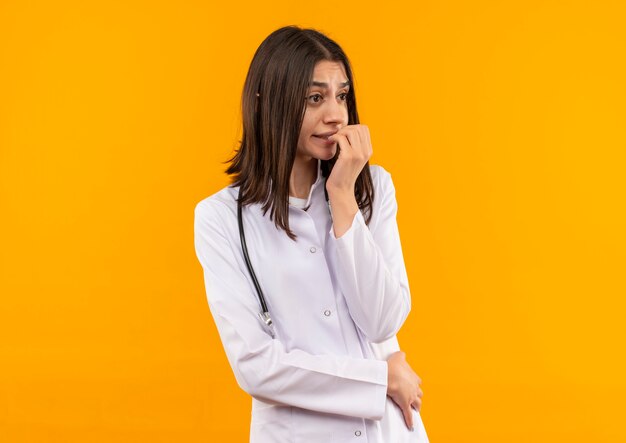 Giovane medico femminile in camice bianco con lo stetoscopio intorno al collo che osserva da parte in piedi stressato e nervoso sopra la parete arancione