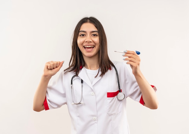 Giovane medico femminile in camice bianco con lo stetoscopio intorno al collo che tiene il termometro digitale che stringe il pugno felice ed eccitato in piedi sopra il muro bianco