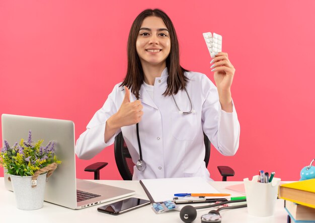 Giovane medico femminile in camice bianco con lo stetoscopio intorno al collo che tiene la bolla con le pillole che mostrano i pollici in su sorridente che si siede al tavolo con il computer portatile sopra la parete rosa